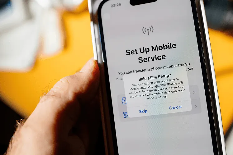 An iPhone 14 Pro rests in a user’s hand, its screen displaying the setup interface with a prompt asking whether to set up an eSIM now or skip this step