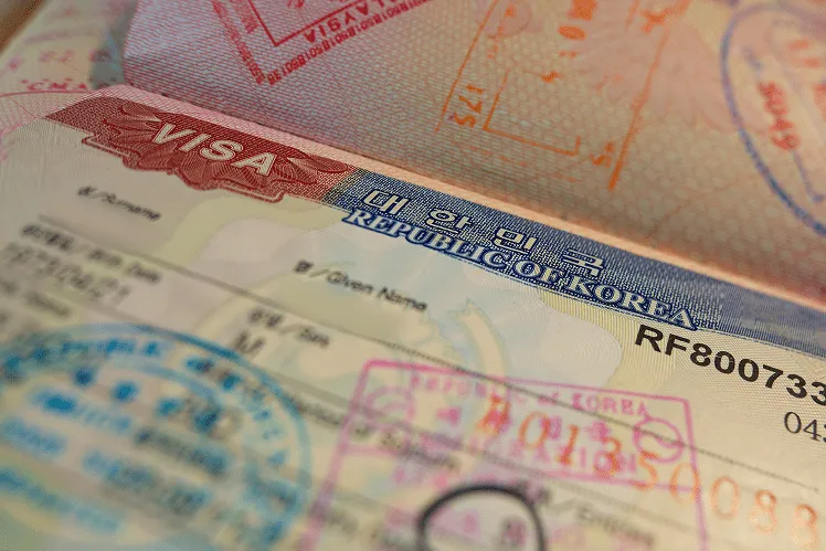 A close-up view captures a Republic of Korea visa page in an open passport, prominently featuring the official entry stamp. The visa displays detailed text in Korean and English
